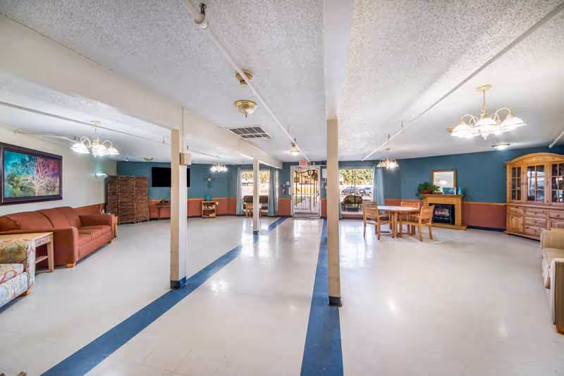 A spacious common area in a senior living facility featuring multiple seating arrangements including sofas and chairs, a wooden dining table with chairs, a wooden cabinet, and a fireplace. The room has blue and white walls, a tiled floor with blue stripes, ceiling lights, and large windows letting in natural light.