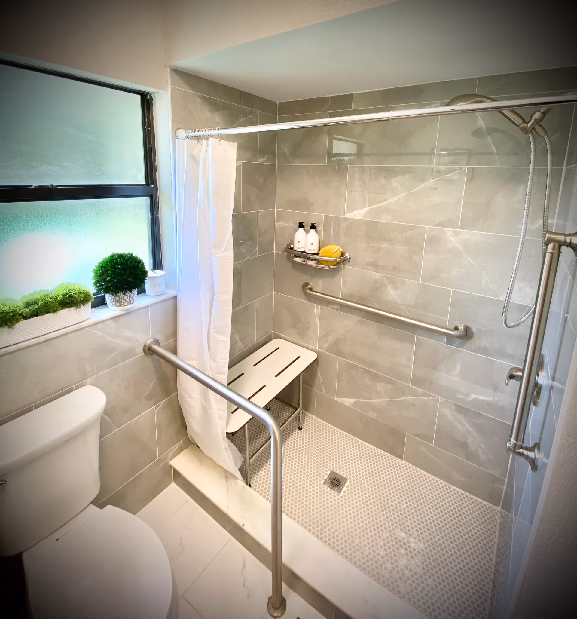 A bathroom with a toilet and a walk-in shower featuring gray tiled walls and a white tiled floor. The shower has a white bench, grab bars, a handheld showerhead, and a small shelf with toiletries. A frosted window with potted plants on the sill provides natural light. A white shower curtain hangs from a curved rod.