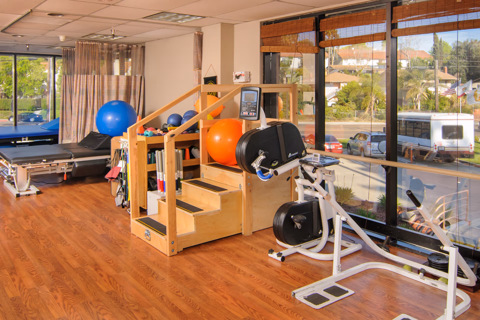 A bright therapy gym with exercise equipment, therapy stairs, and large windows overlooking the street.