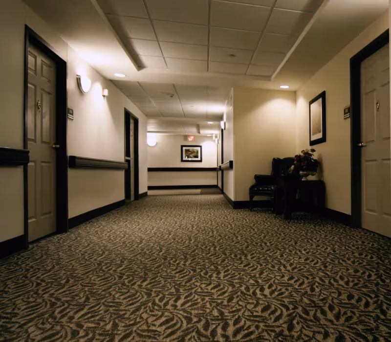 Carpeted interior hallway with doors on both sides, wall sconces, framed artwork, and a chair with a side table and flowers.