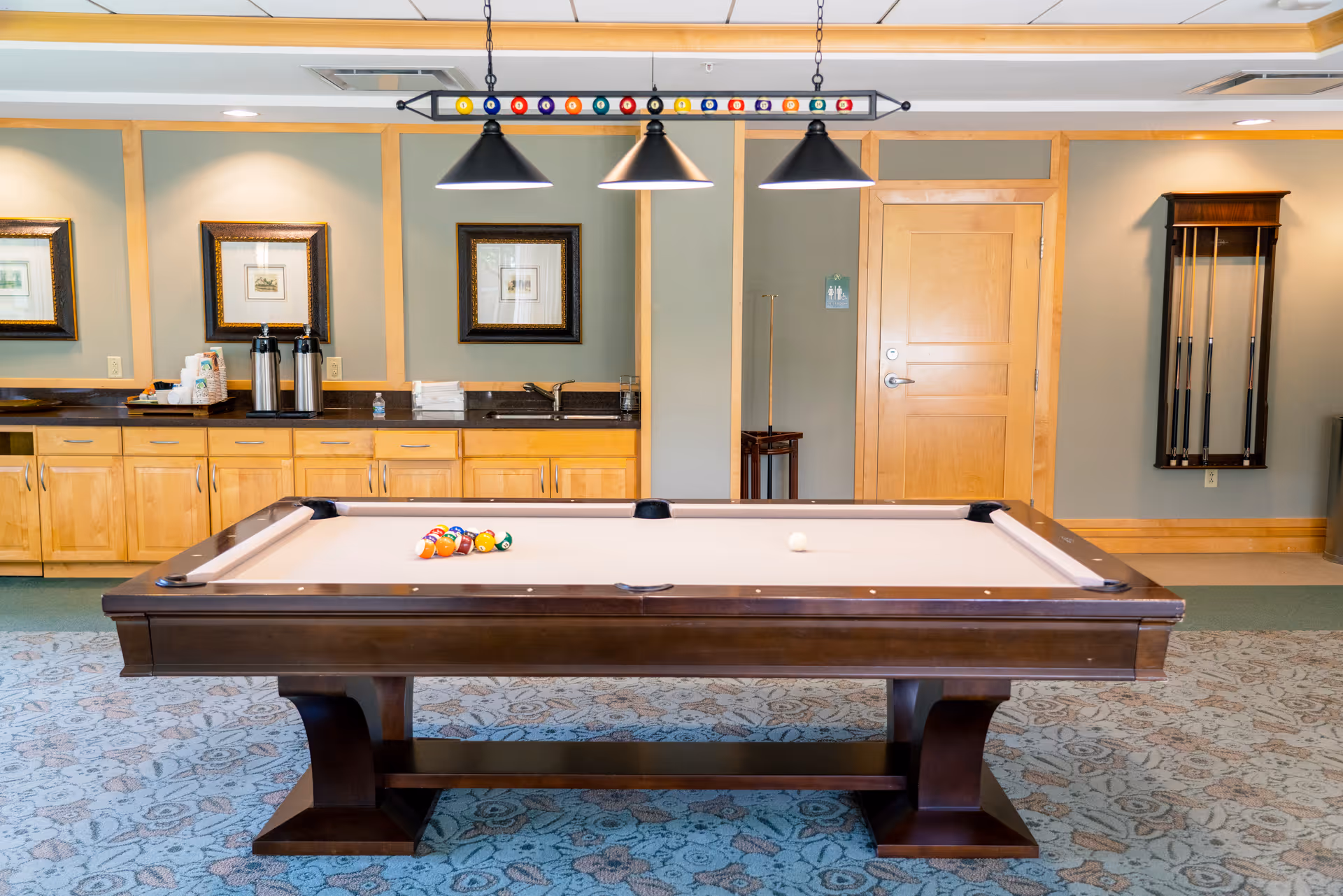 A well-lit game room featuring a pool table with a rack of billiard balls arranged for a game. Above the table hang three black pendant lights with a decorative billiard ball rack. The background shows wooden cabinets with a countertop holding coffee dispensers and supplies, framed artwork on the wall, a wooden door, and a wall-mounted cue rack with pool cues.