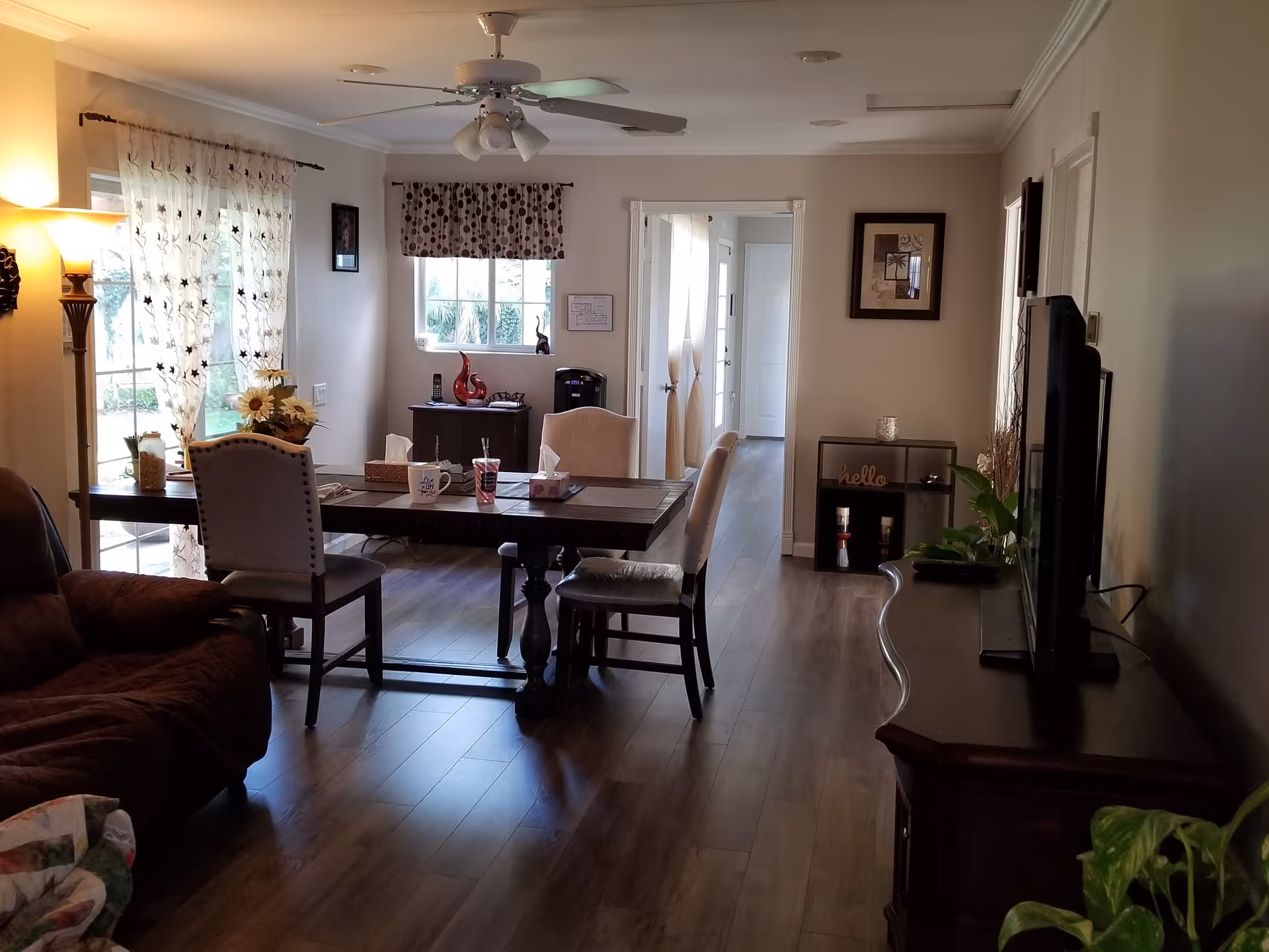 A cozy living and dining area with wooden flooring, a ceiling fan with lights, a dining table with chairs, a brown recliner chair, a TV on a wooden stand, and decorative items including plants, framed pictures, and curtains on the windows.