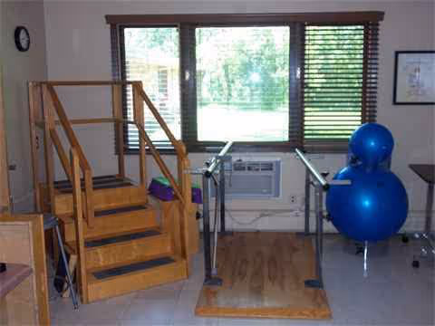 Interior therapy room with parallel bars, wooden stairs, and blue exercise balls by a window.