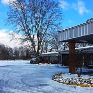 Snow-covered parking area and building entrance with a large tree beside a brick-columned entry.