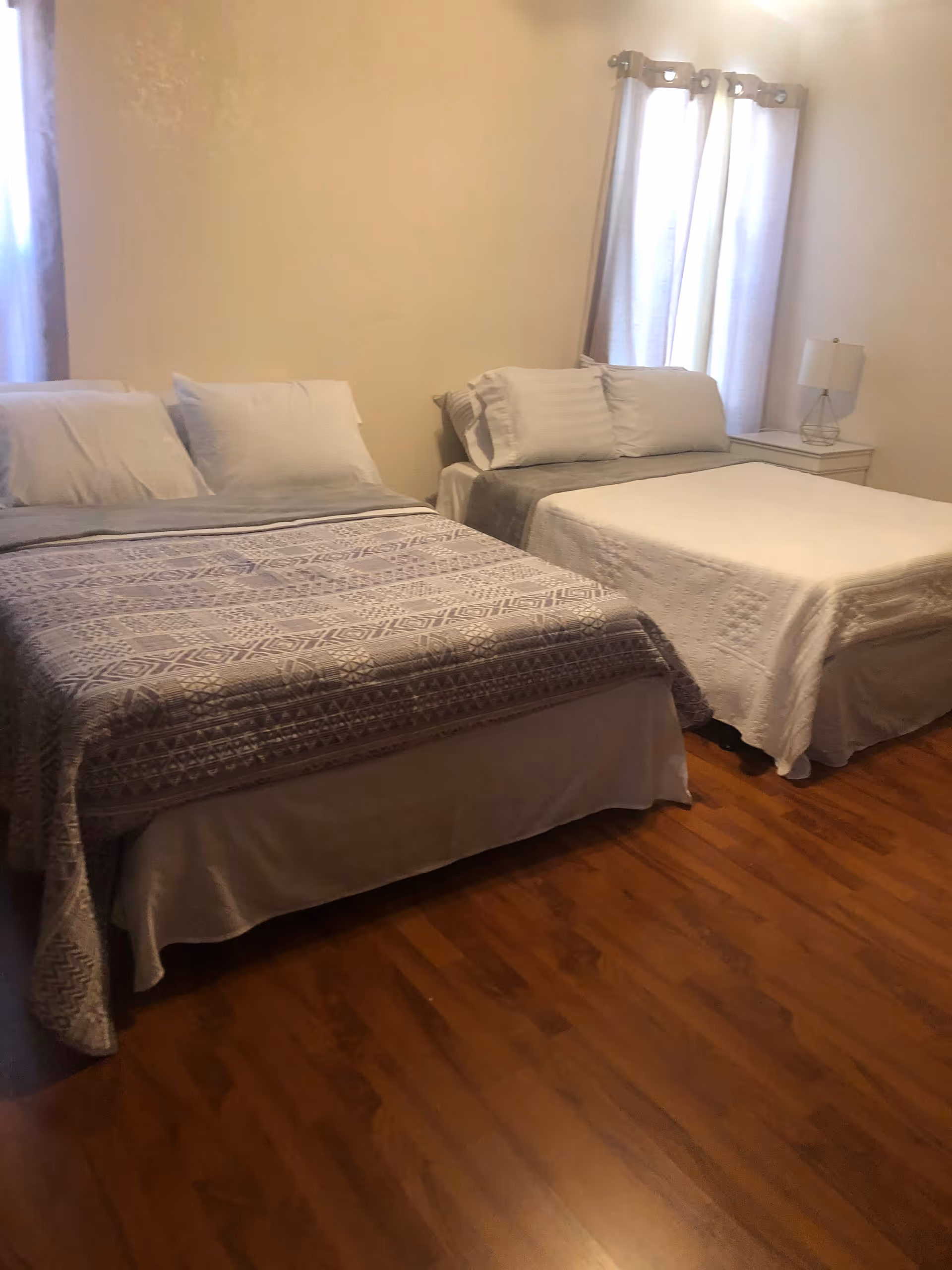 A bedroom with two beds side by side, each made with white pillows and blankets. The bed on the left has a patterned gray and white blanket, while the bed on the right has a plain white blanket. There is a small white nightstand between the beds with a lamp on it. The room has wooden flooring and light-colored walls with two windows covered by light curtains.
