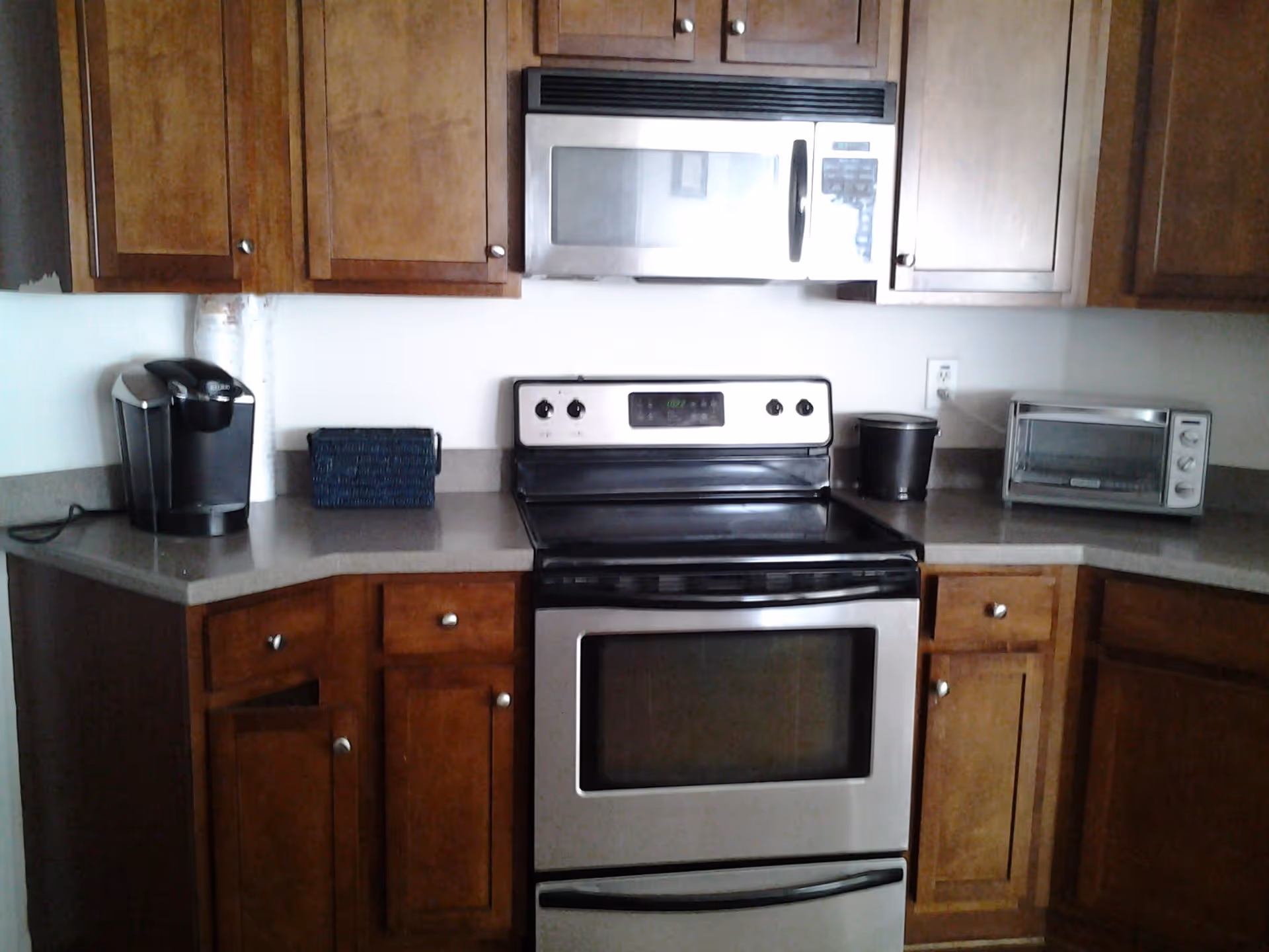 A kitchen area with wooden cabinets, a stainless steel microwave mounted above a stainless steel electric stove and oven. On the countertop to the left is a black coffee maker and a small blue basket. To the right on the countertop is a black container and a small silver toaster oven.