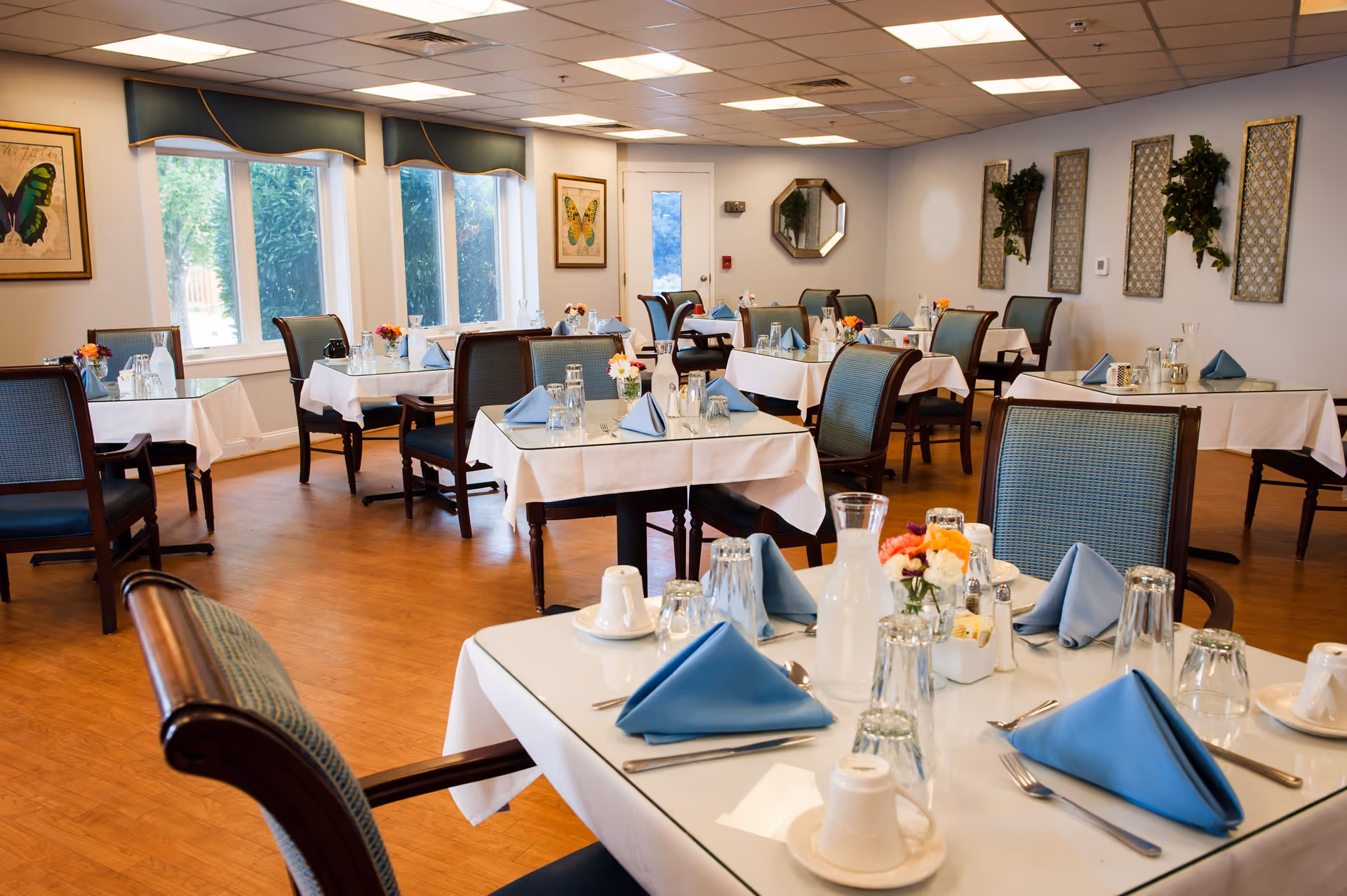 A bright dining room with multiple tables covered in white tablecloths and set with blue folded napkins, glasses, silverware, and small flower arrangements. The room has large windows with green valances, butterfly artwork on the walls, and wooden flooring.