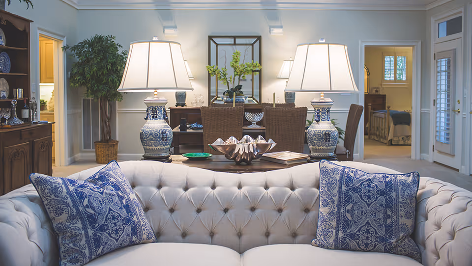 A cozy living room area with a tufted beige sofa adorned with two blue and white patterned pillows. Behind the sofa, there is a wooden table with two large blue and white ceramic lamps, a decorative bowl, and a book. The room features a wooden cabinet with glassware on the left, a potted plant, and a mirror on the back wall reflecting the lamps. Doorways lead to other rooms, including a bedroom visible on the right side.