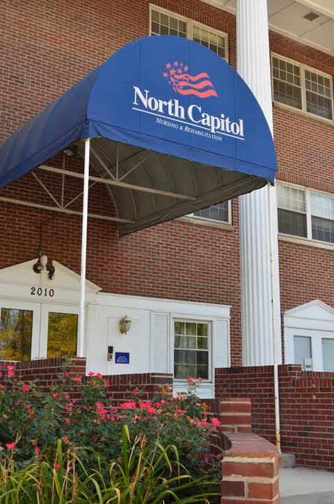 Entrance of North Capitol Nursing and Rehabilitation facility with a blue awning displaying the facility's name and logo, brick exterior walls, white framed windows, and a garden with pink flowers in front.