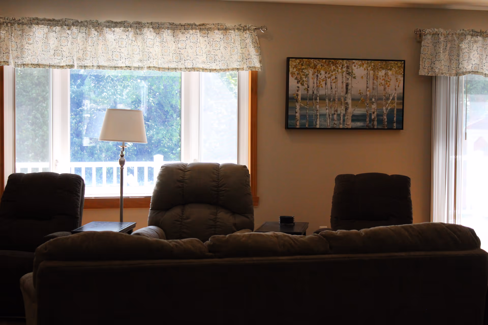 A cozy living room with a large window covered by a floral valance, a floor lamp in front of the window, three cushioned armchairs, a cushioned sofa facing away from the camera, two small side tables, and a framed painting of birch trees on the wall.