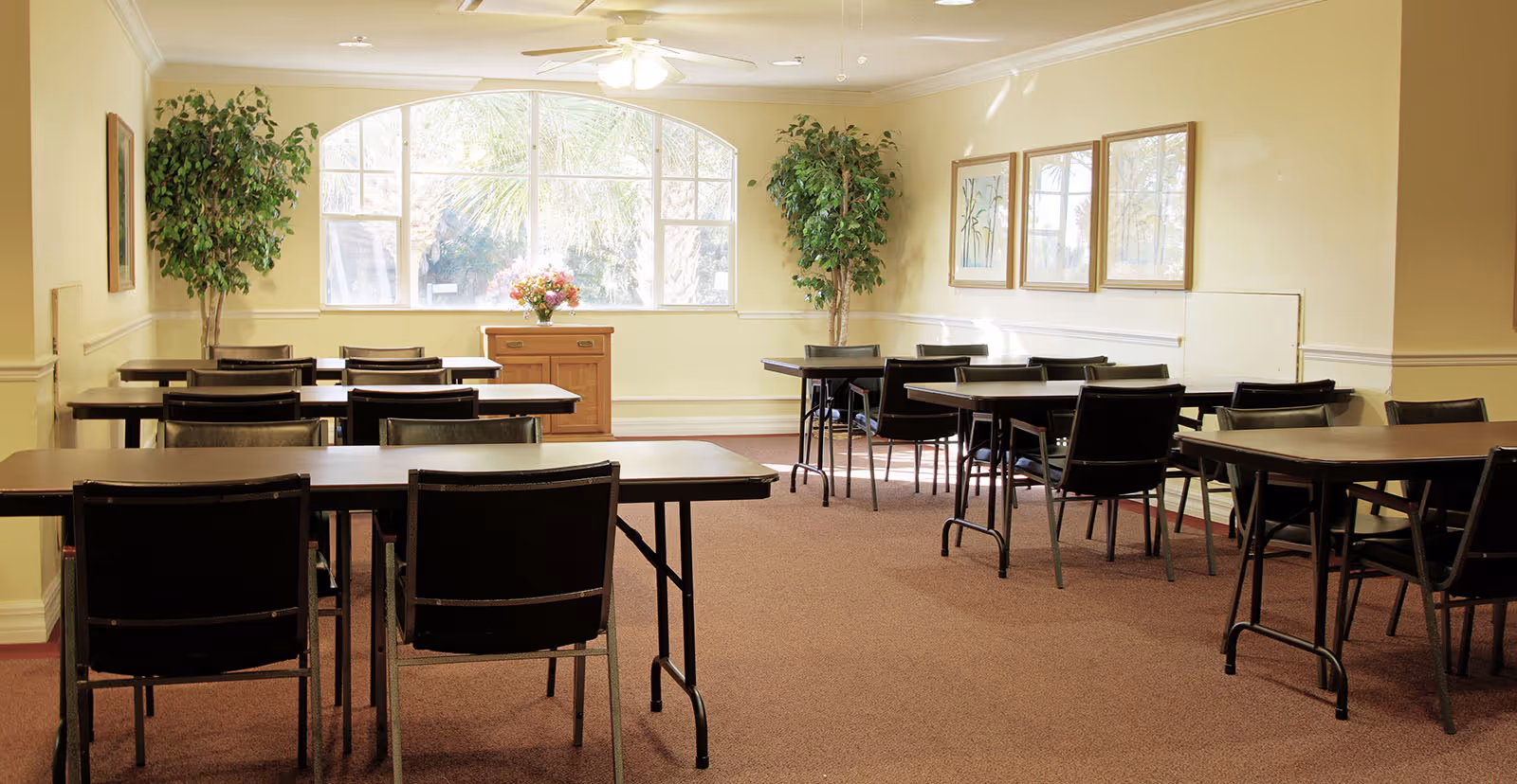 Bright multipurpose room with rows of tables and chairs, potted plants, and a large arched window.