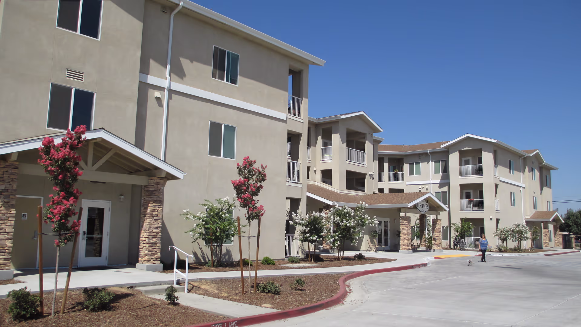 Three-story beige retirement community building with covered entrances, small landscaped trees, and a paved driveway under a clear blue sky.