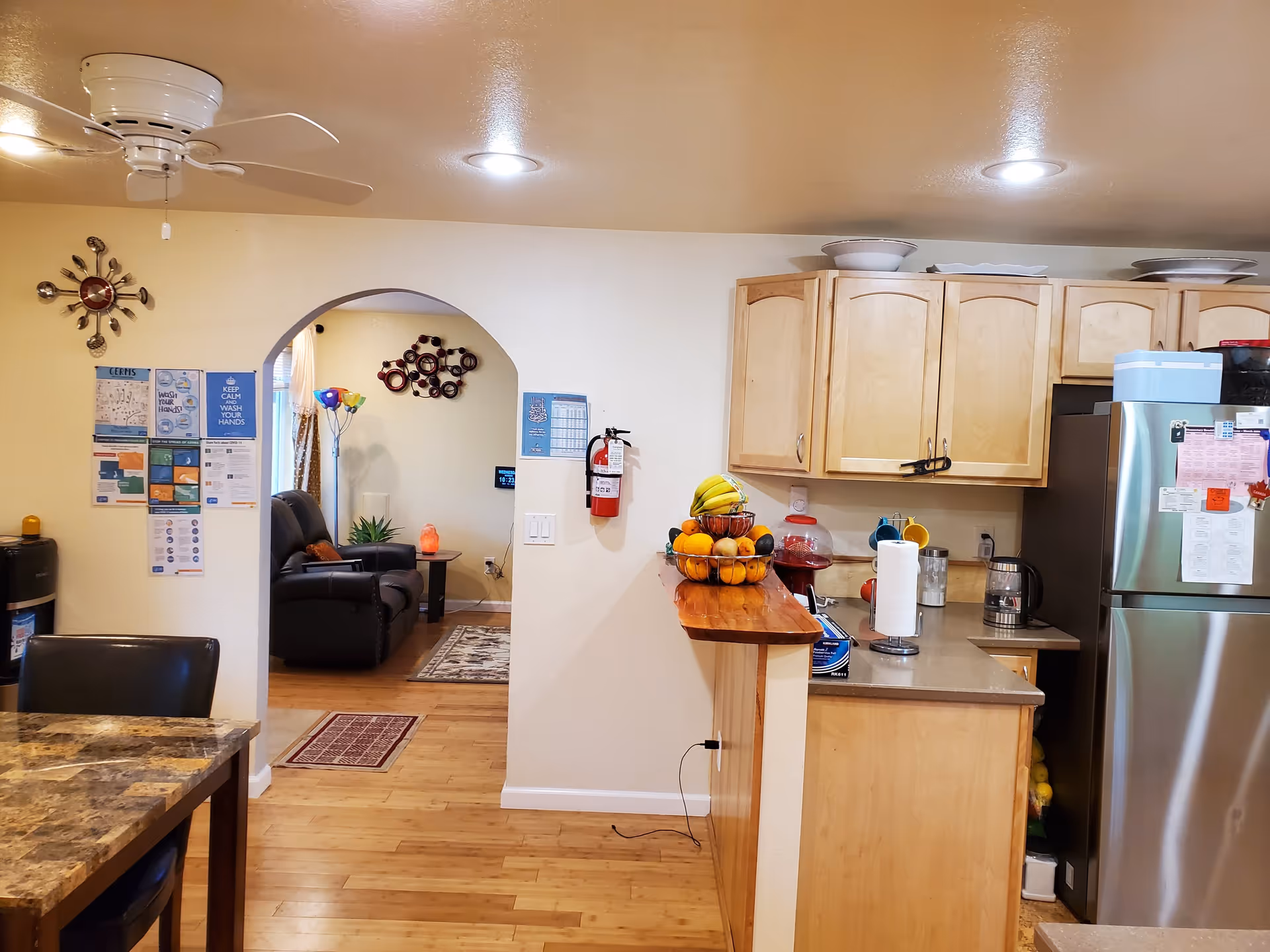 Open kitchen with light wood cabinets, stainless steel refrigerator and a countertop fruit bowl, looking through an arched doorway into a living room.