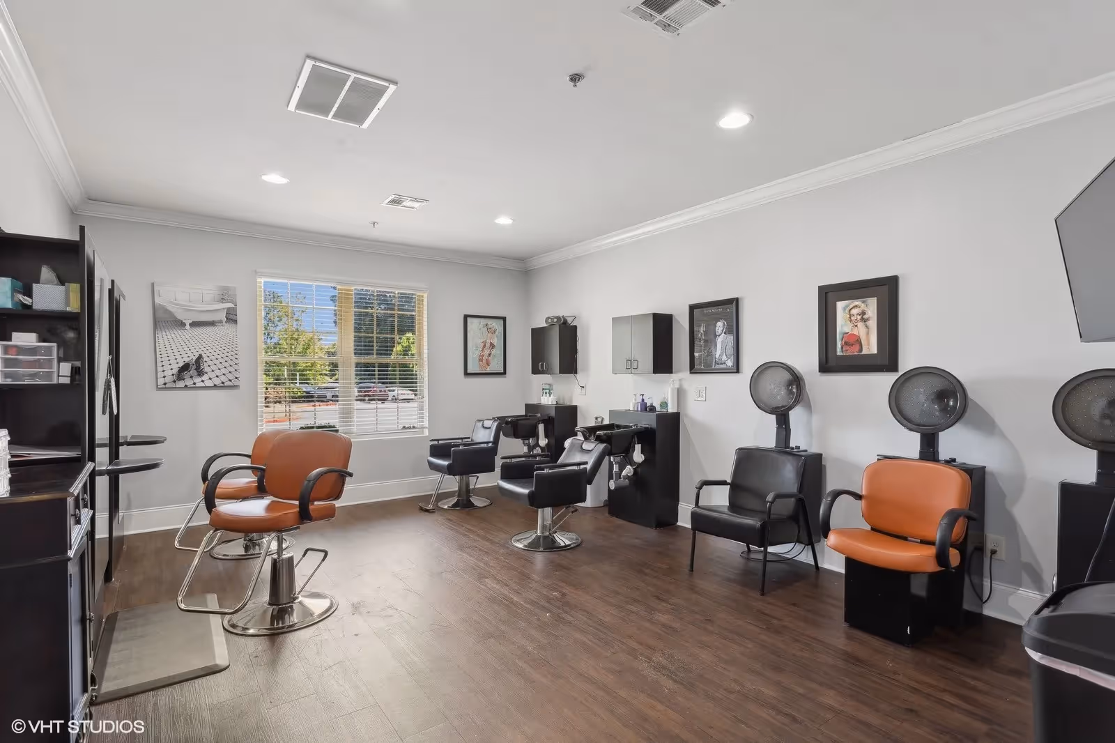 Interior view of a hair salon with multiple styling chairs, hair washing stations, and hair dryers. The room has wooden flooring, white walls with framed pictures, and a large window letting in natural light.