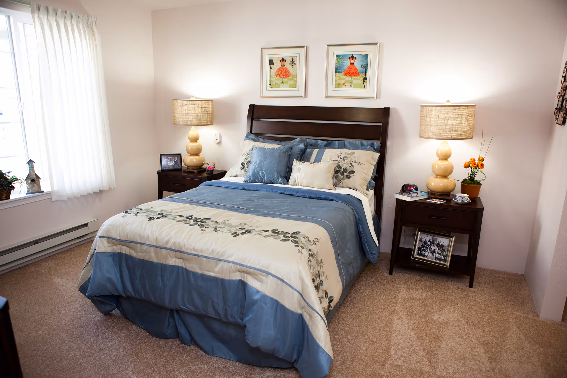 Sunlit bedroom with a blue-and-cream bed, wooden headboard, matching nightstands with lamps and decor, and a window with curtains.