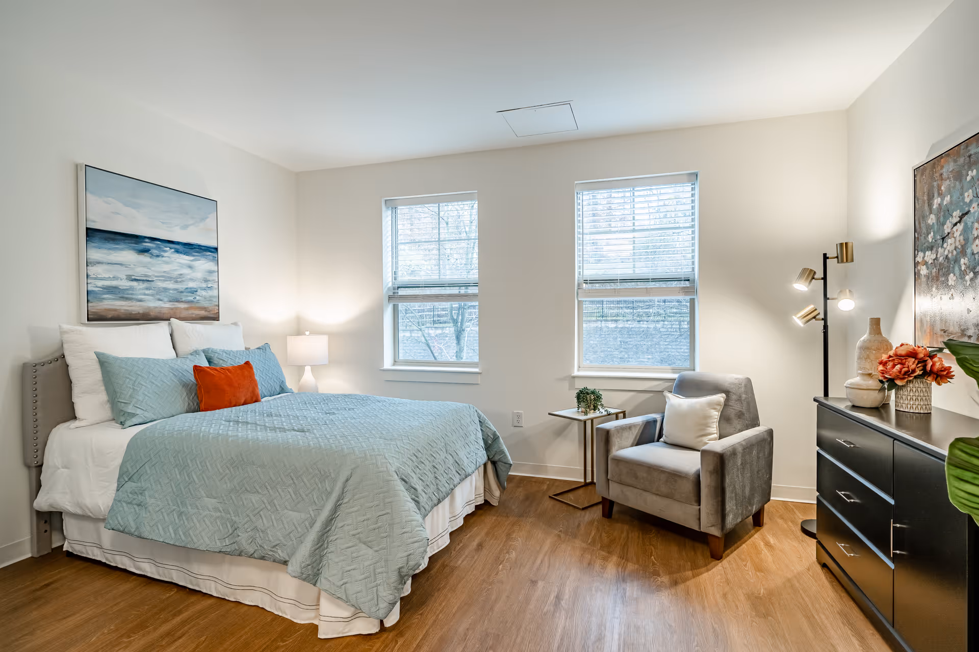 A bright and cozy bedroom with a neatly made bed featuring light blue bedding and an orange accent pillow. There is a gray armchair with a white pillow next to a small side table with a plant. The room has two windows with blinds, a black dresser with decorative items, a floor lamp, and a large painting on the wall above the bed and another above the dresser. The floor is wooden, and the walls are painted white.