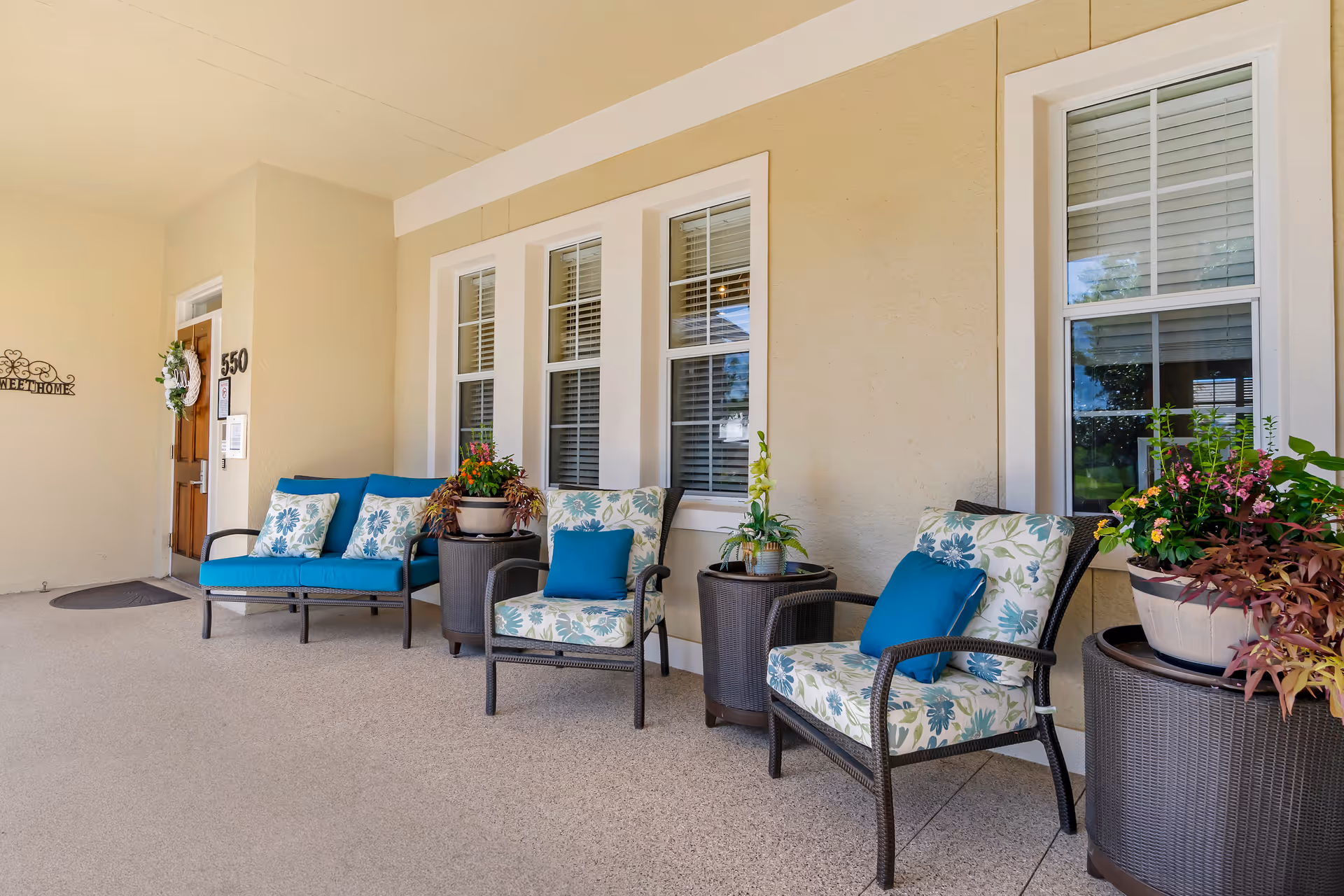 Covered front porch with wicker chairs and a loveseat with blue cushions and potted plants outside a building entrance.
