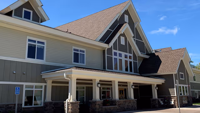Exterior view of a large, two-story building with beige and gray siding, multiple windows, and a covered entrance supported by columns. The building has a steeply pitched roof with dormer windows and stone accents at the base. A handicap parking sign is visible near the entrance under a clear blue sky.