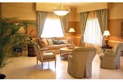 Sunlit communal living room with a sofa and armchairs arranged around a coffee table, lamps, potted plants, and curtained windows.