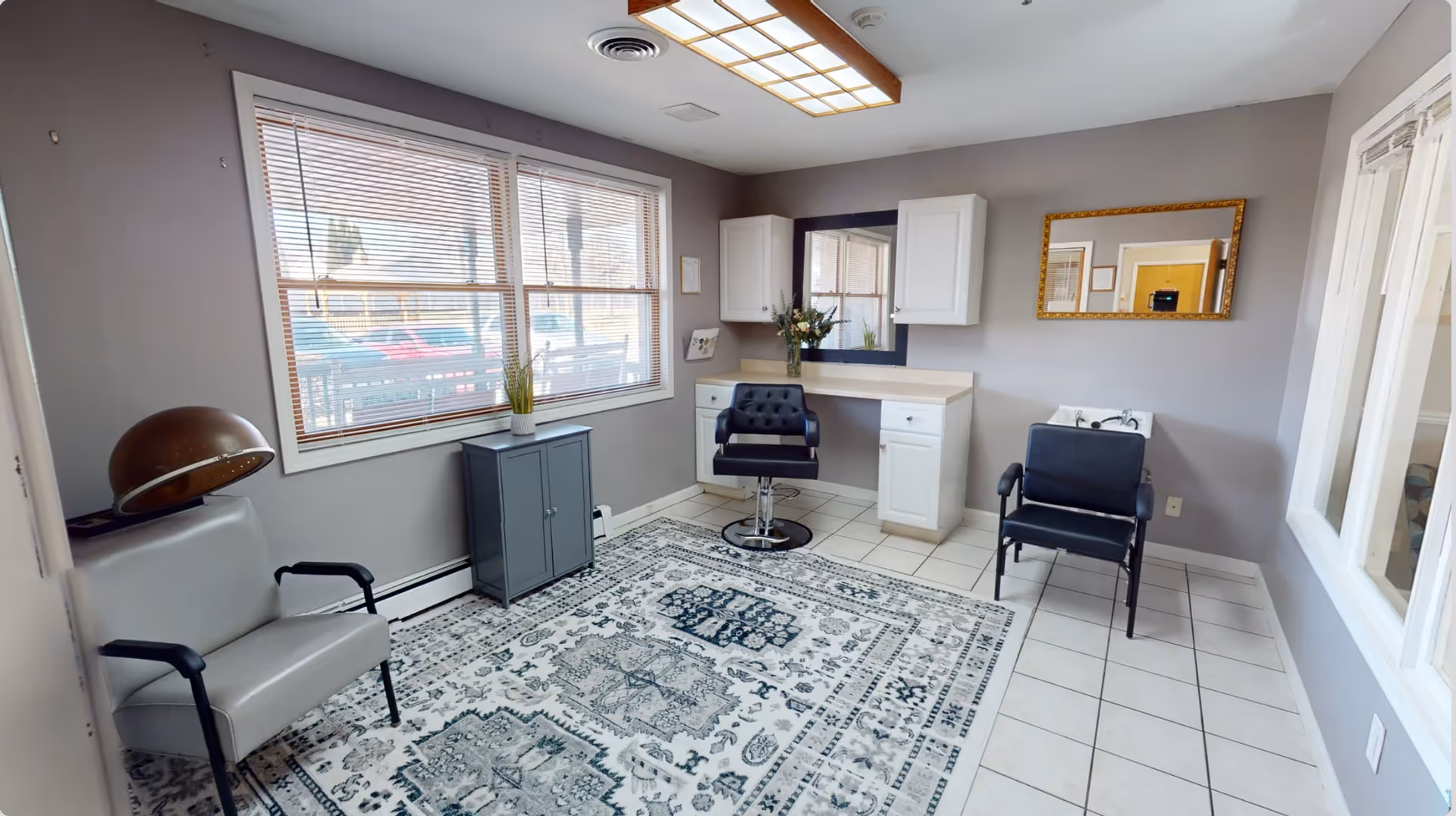A small salon room with a hair dryer chair on the left, a styling chair in front of a mirror and counter with white cabinets, and another chair next to a sink. The room has a large window with blinds, a patterned area rug on tiled floor, and light gray walls with a framed mirror.