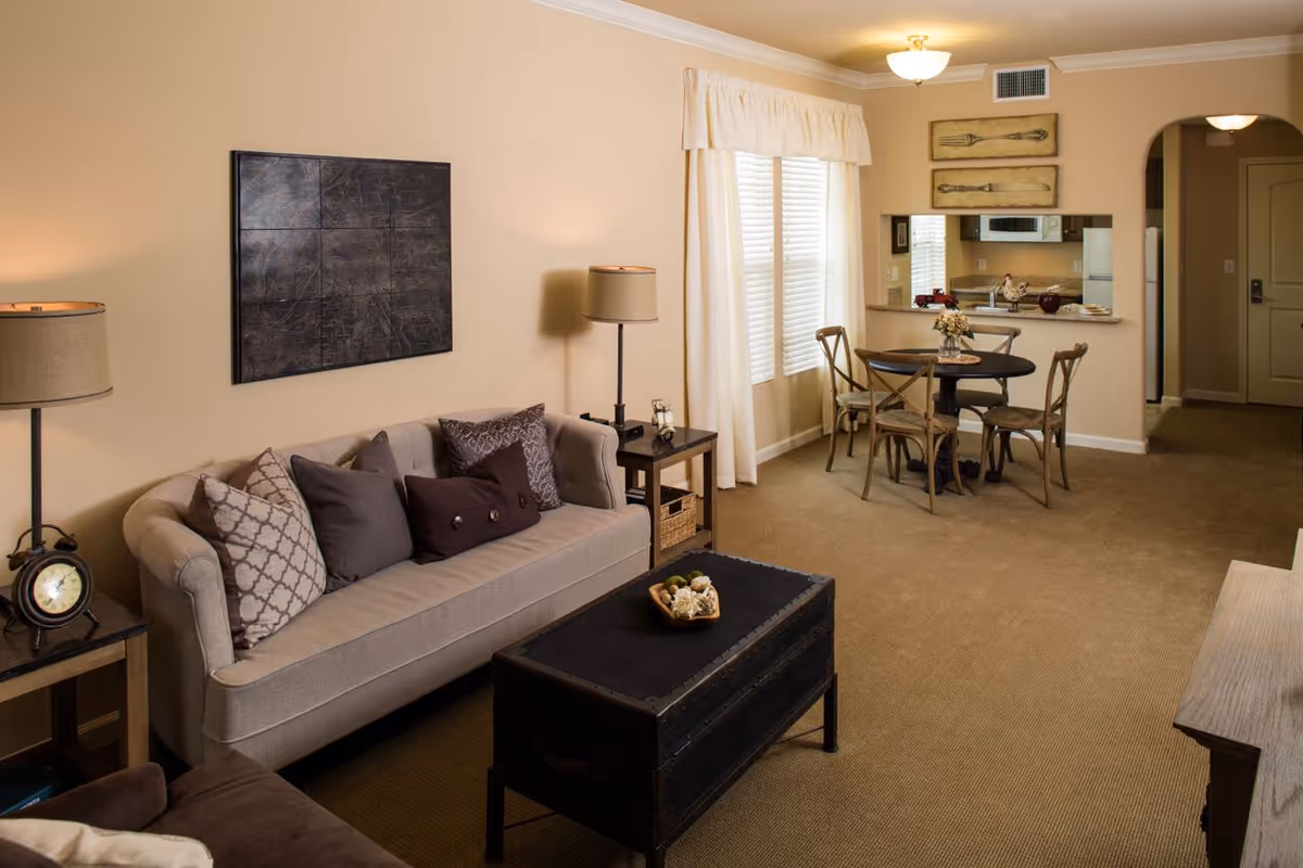 A cozy living room area with a beige sofa adorned with multiple decorative pillows, two side tables with lamps, a black trunk-style coffee table with a decorative bowl on top, and a small round dining table with four wooden chairs near a window with white curtains. The room has beige walls and carpet, with a kitchen visible through a pass-through window in the background.