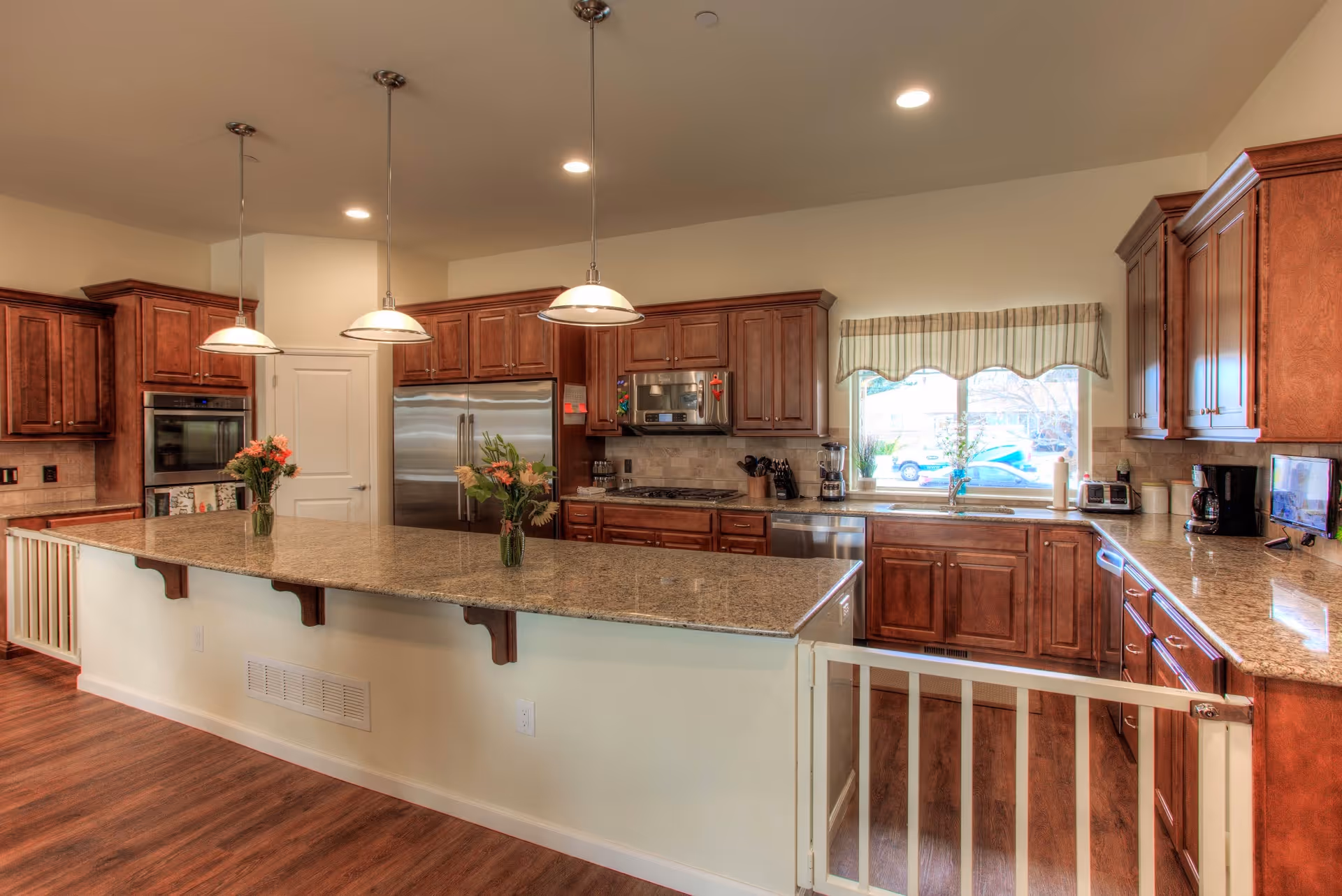 Bright kitchen with a long granite island, pendant lights, wooden cabinets and stainless steel appliances.