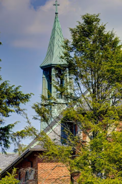 A brick building with a green copper steeple topped by a cross, partially obscured by trees.