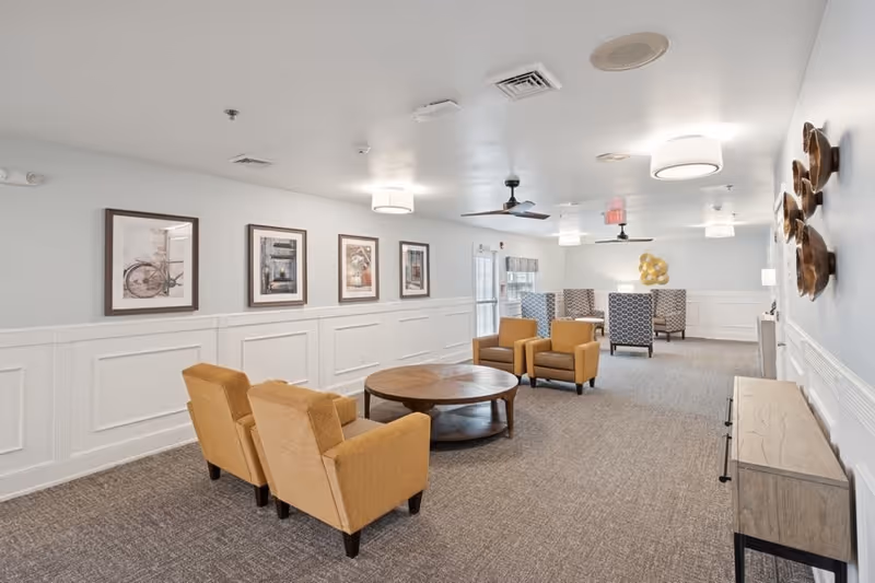 A spacious, well-lit common area with beige armchairs and a round wooden coffee table in the foreground. The walls are painted light gray with white wainscoting and decorated with framed pictures and wall art. The room has ceiling fans, modern light fixtures, and carpeted flooring. In the background, there are additional seating areas with patterned chairs and a door leading outside.