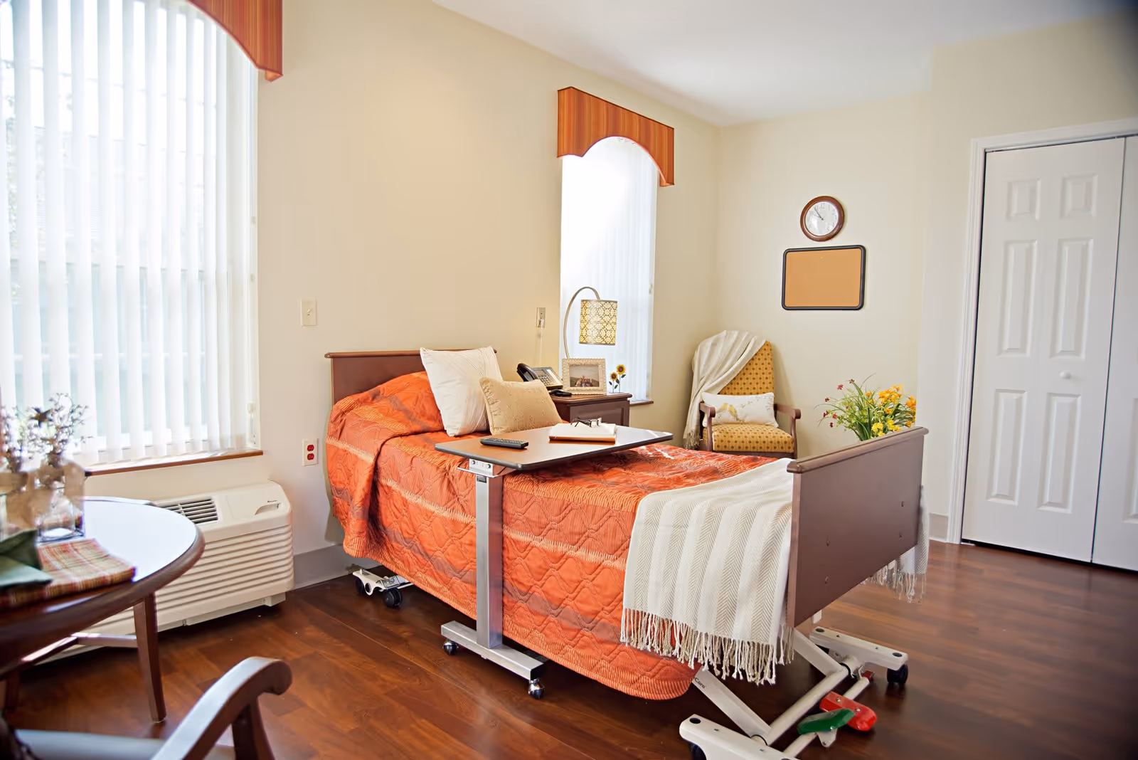 A bright and clean senior living bedroom with a hospital-style bed covered in an orange quilt and a white throw blanket. The room has two tall windows with vertical blinds and orange valances. There is a small table with a lamp, phone, and picture frame next to the bed. A yellow armchair with a white pillow and a blanket draped over it sits in the corner. A round table with flowers and a chair are partially visible. The floor is wooden, and there is a white closet door on the right wall.