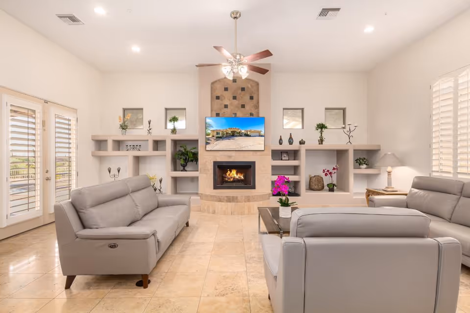 A bright and spacious living room with beige tiled floors, light gray leather sofas arranged around a glass coffee table with a pink orchid plant. The room features a central fireplace with a mounted flat-screen TV above it, built-in shelves with decorative items, and large windows with white shutters allowing natural light to fill the space. A ceiling fan with lights hangs from the ceiling.