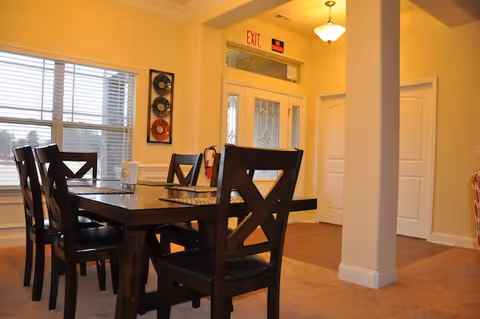 Interior view of a dining area in Avendelle Assisted Living featuring a dark wooden dining table with six matching chairs. The room has beige walls, a window with blinds, a decorative wall piece, and a visible entrance door with an exit sign above it.