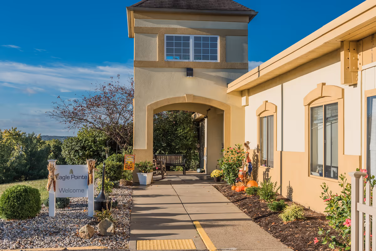 Front entrance walkway and arched entry of Eagle Pointe Healthcare Center with landscaped beds, a welcome sign, and seasonal pumpkins.