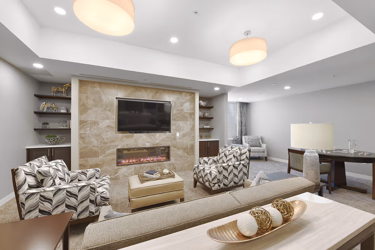 A modern communal living room with patterned armchairs, a sofa, a wall-mounted TV above a fireplace, shelving, and table lamps.