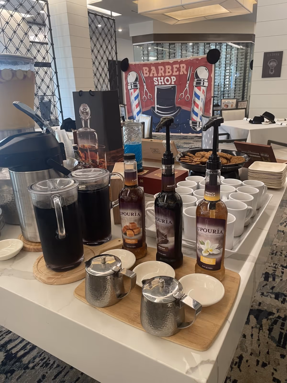 A coffee and beverage station with syrup bottles labeled Caramel, Mocha, and French Vanilla, two pitchers filled with dark liquid, two metal creamers, stacks of white cups, and a plate of cookies. In the background, there is a sign that reads 'Barber Shop' with barber poles and scissors illustrations, and tables with chairs in a well-lit room.