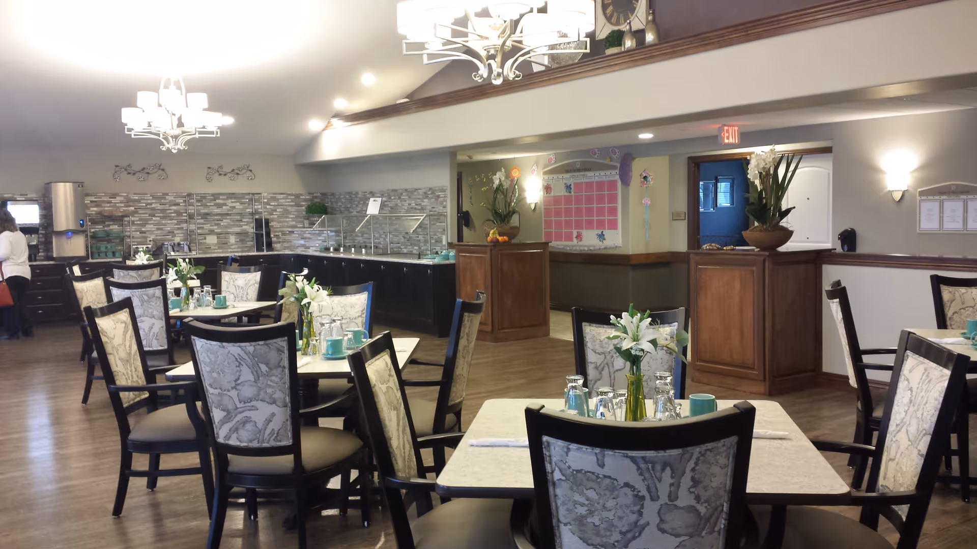 Dining room with multiple set tables and patterned chairs, a buffet counter in the background and decorative chandeliers.