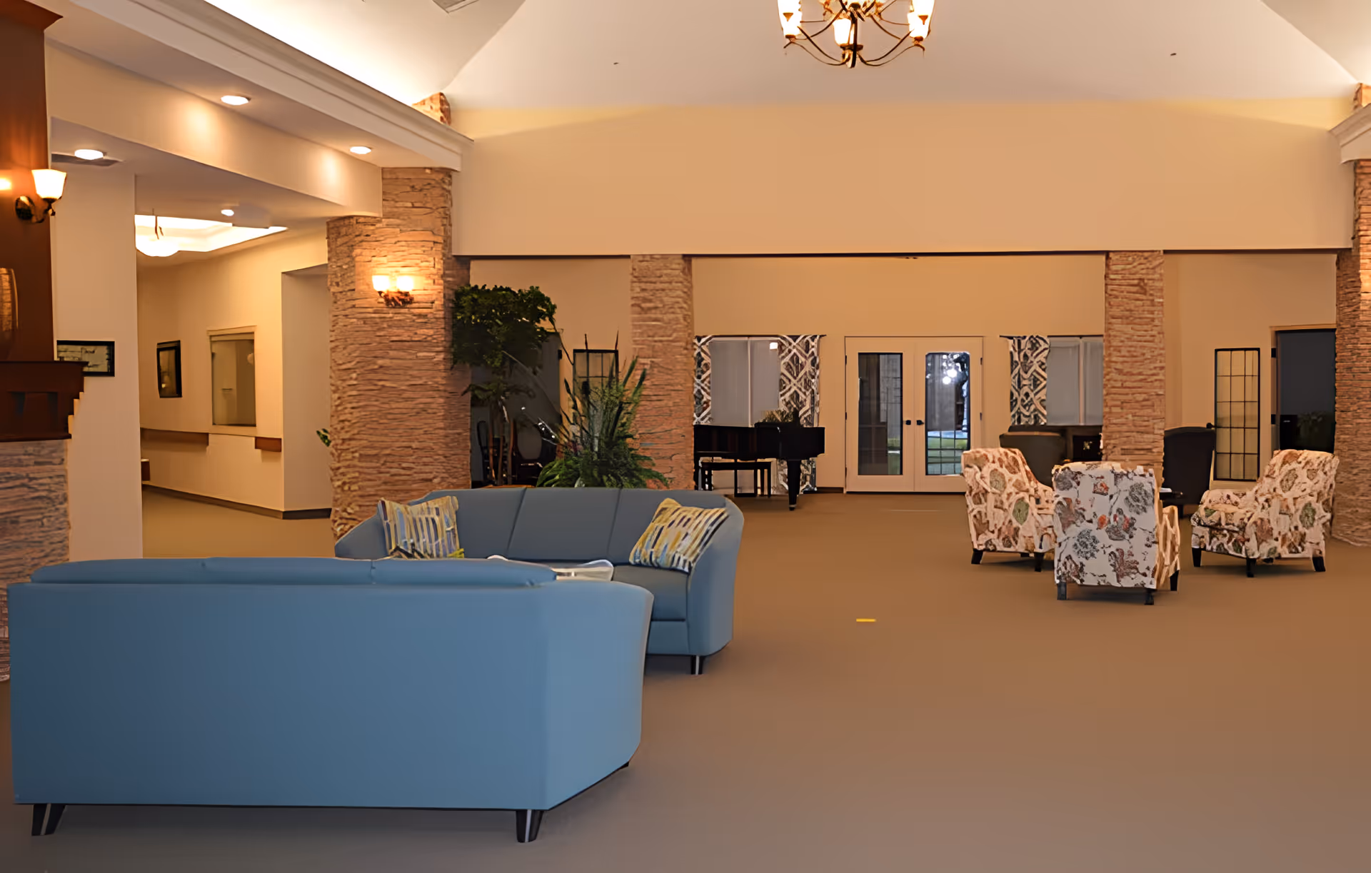Spacious senior living facility common area with blue sofas and floral armchairs arranged around a coffee table. The room features stone pillars, warm lighting, a chandelier, and a black grand piano near glass doors with patterned curtains.