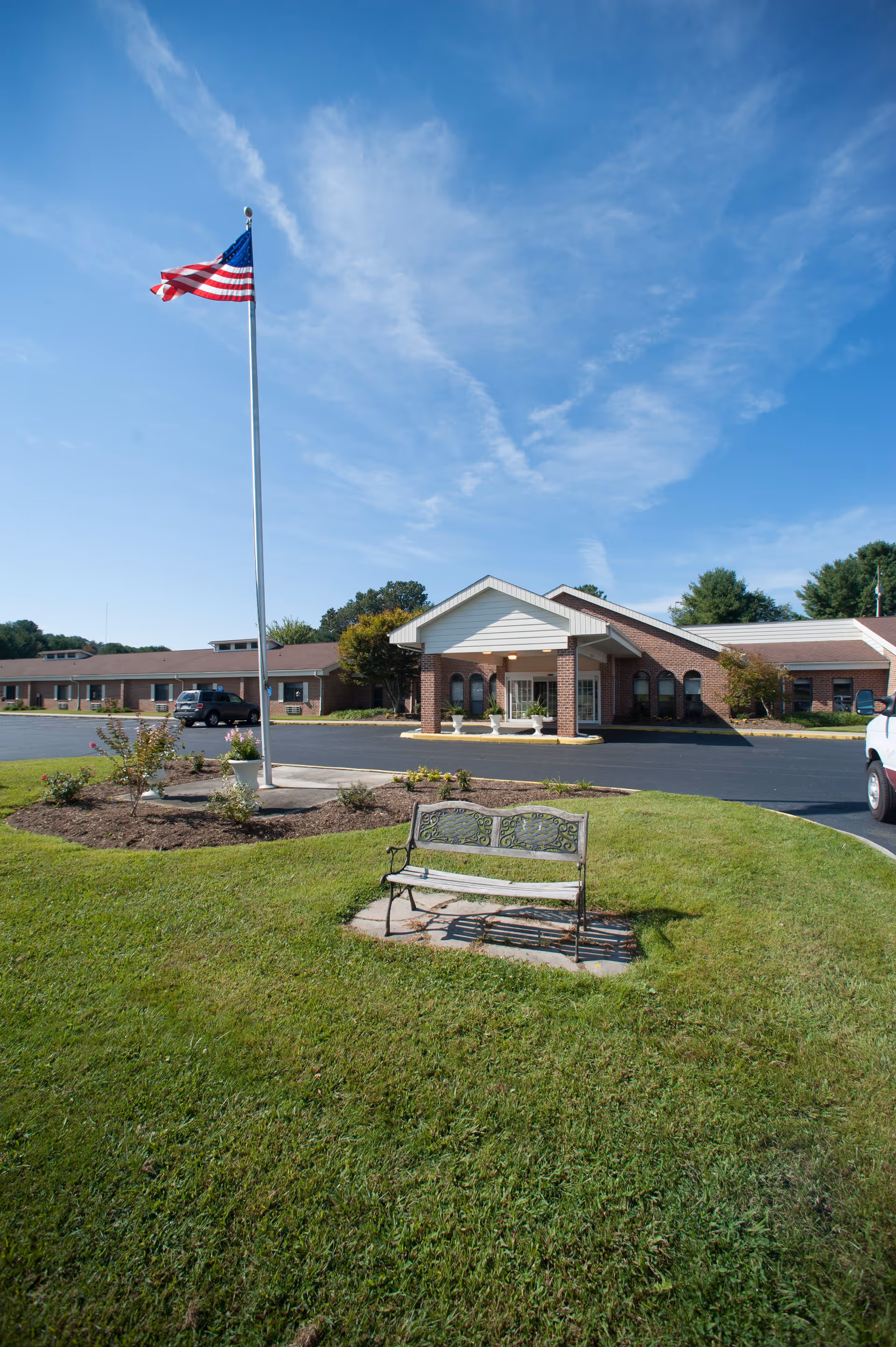 Exterior view of Commonwealth Senior Living At Hillsville showing a single-story brick building with a covered entrance, an American flag on a tall flagpole, a bench on a grassy area, and a clear blue sky.