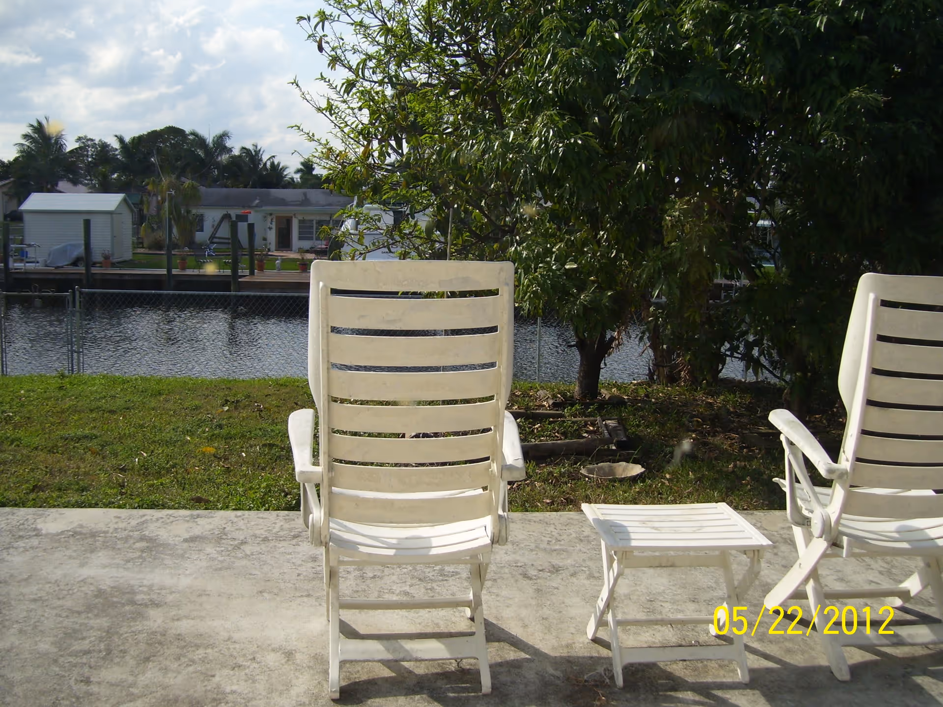 Two white plastic lounge chairs and a small matching table on a concrete patio facing a grassy area with a tree and a canal with houses and docks on the opposite side under a partly cloudy sky.