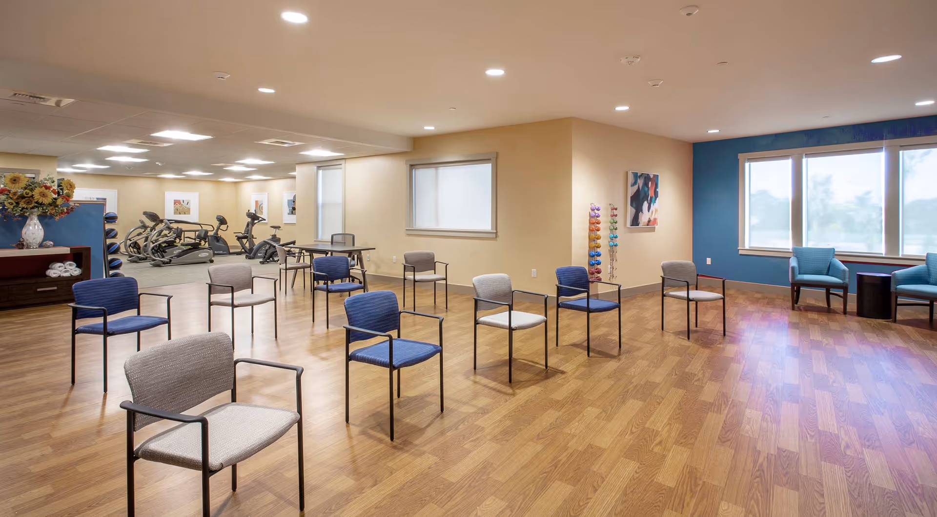 A spacious room with wooden flooring featuring several chairs arranged in a semi-circle, a rack of colorful dumbbells, and exercise equipment such as stationary bikes in the background. The room has large windows with blinds, light-colored walls, and ceiling lights providing bright illumination.