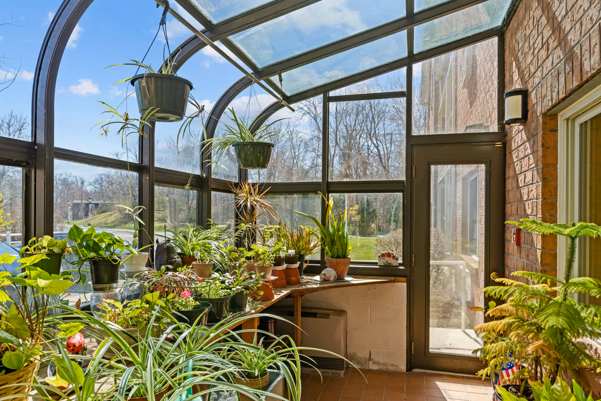 A bright sunroom with large glass windows and a glass ceiling, filled with various potted plants on shelves and hanging from the ceiling. Outside, trees and a grassy area are visible under a clear blue sky. The room has a tiled floor and a door leading outside.