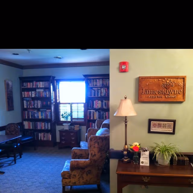 Cozy assisted-living common room with bookshelves, upholstered chairs, a table with a lamp and plants, and a 'Jamestowne Assisted Living' sign on the wall.