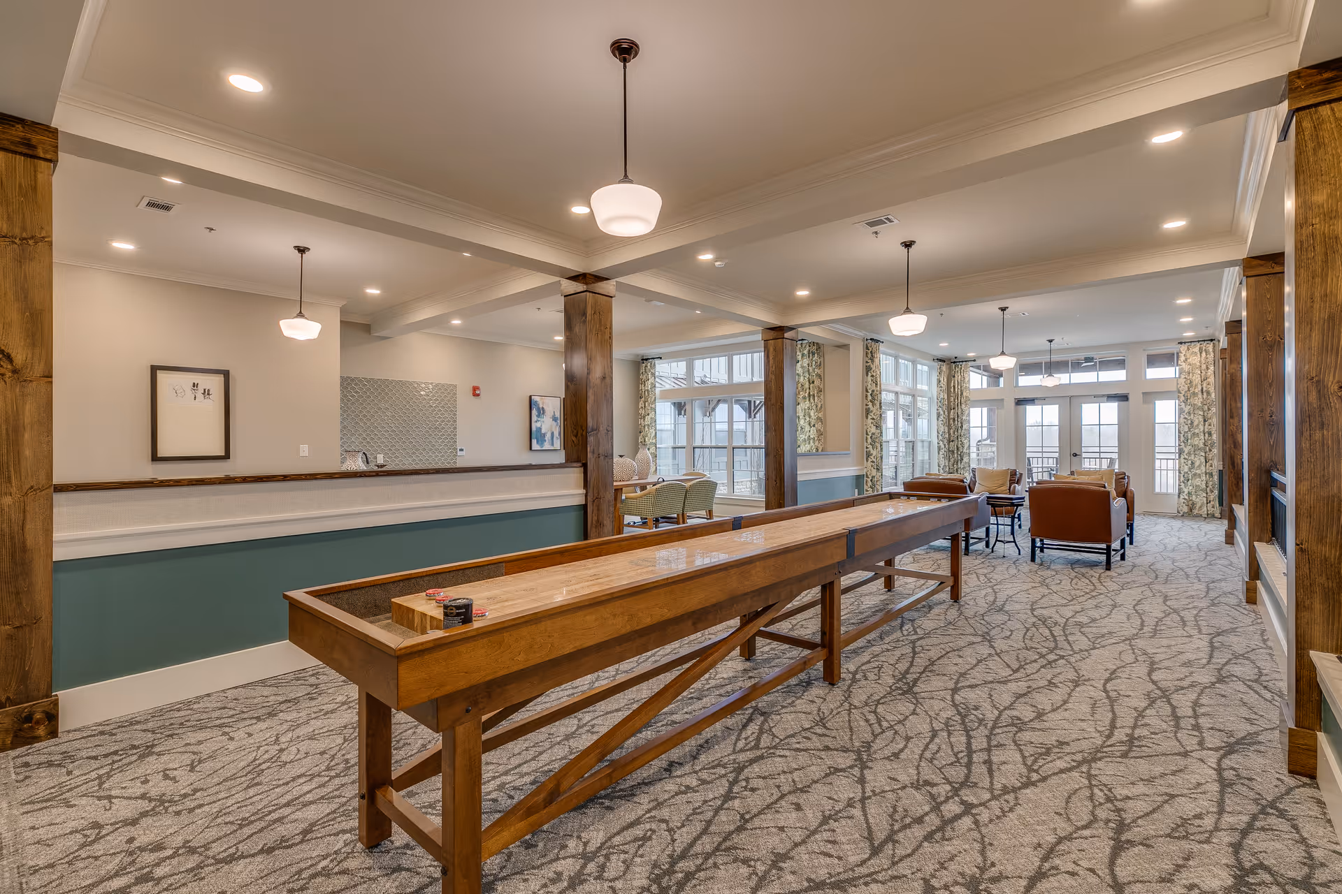 Bright communal lounge with a long wooden shuffleboard table, seating area, and large windows.