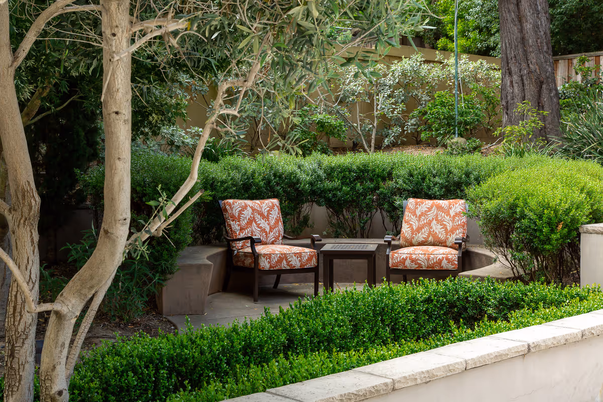 Two patterned cushioned chairs and a small table set in a lush outdoor garden courtyard surrounded by hedges and trees.