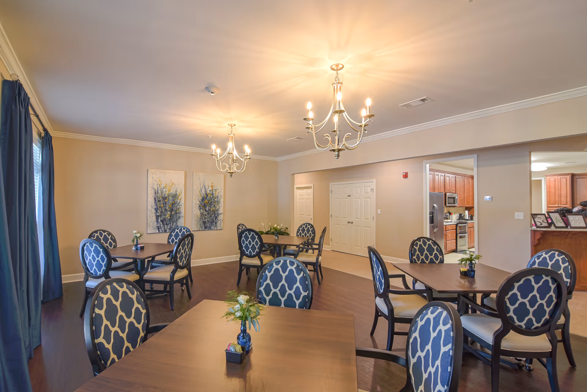 A dining room with several wooden tables and blue patterned chairs. Each table has a small flower arrangement. The room has beige walls, two chandeliers, and two paintings on one wall. There is a view into a kitchen area with wooden cabinets in the background.