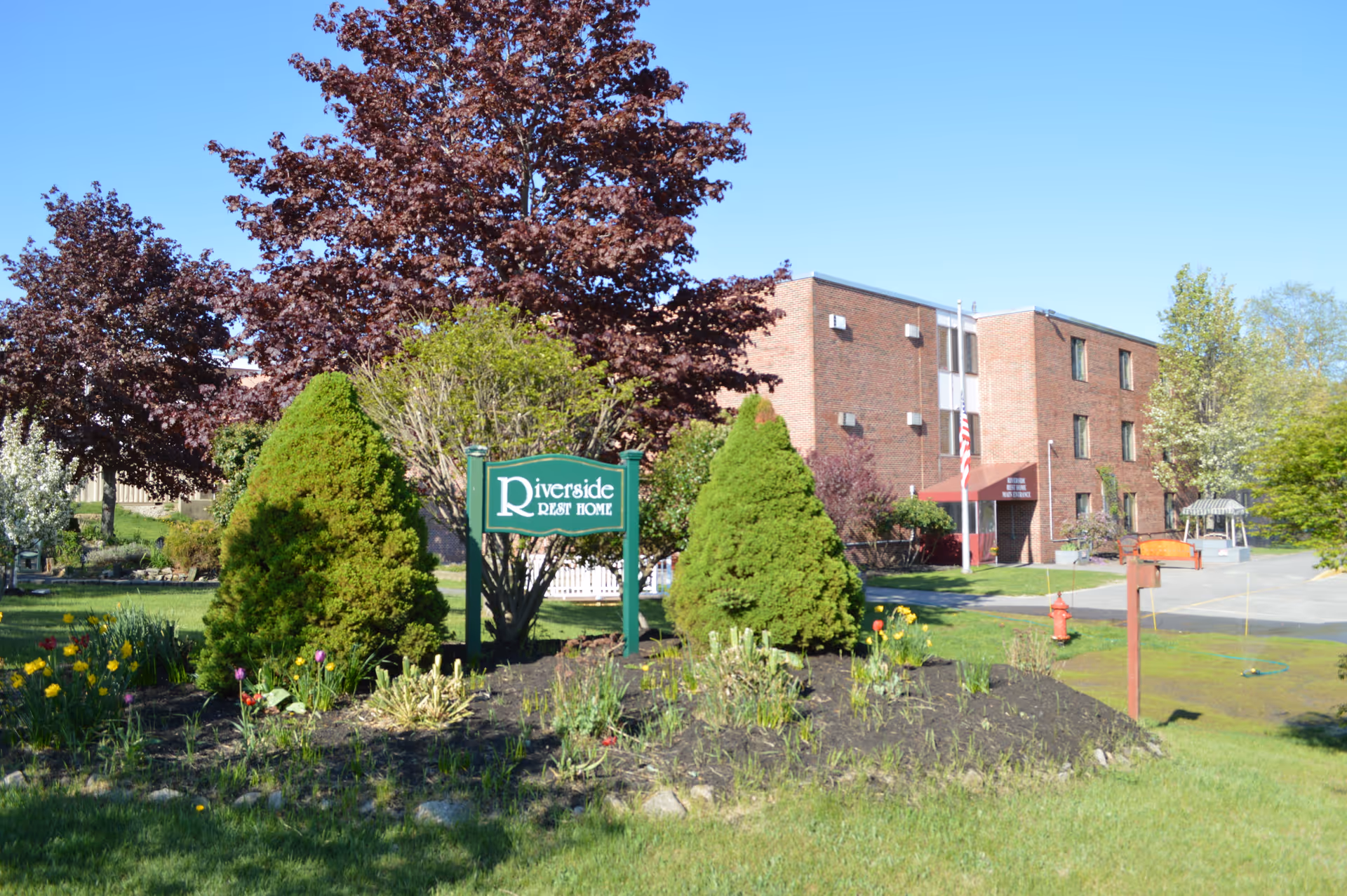 Exterior view of Riverside Rest Home showing a brick building with three floors, surrounded by green trees and bushes. There is a green sign in front with the facility name, and a clear blue sky overhead.