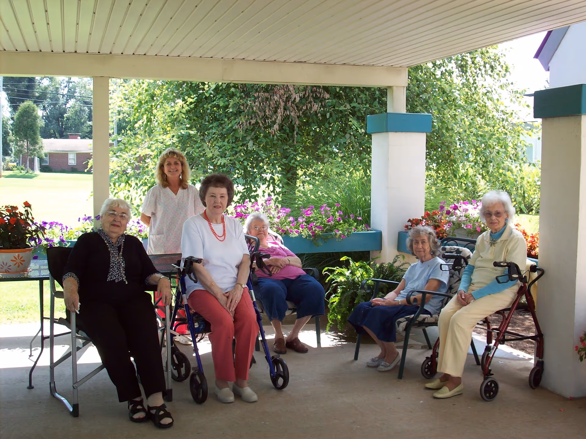 A group of five elderly women and one caregiver sitting and standing under a covered outdoor patio area with flowers and greenery in the background. Two of the women are using walkers.