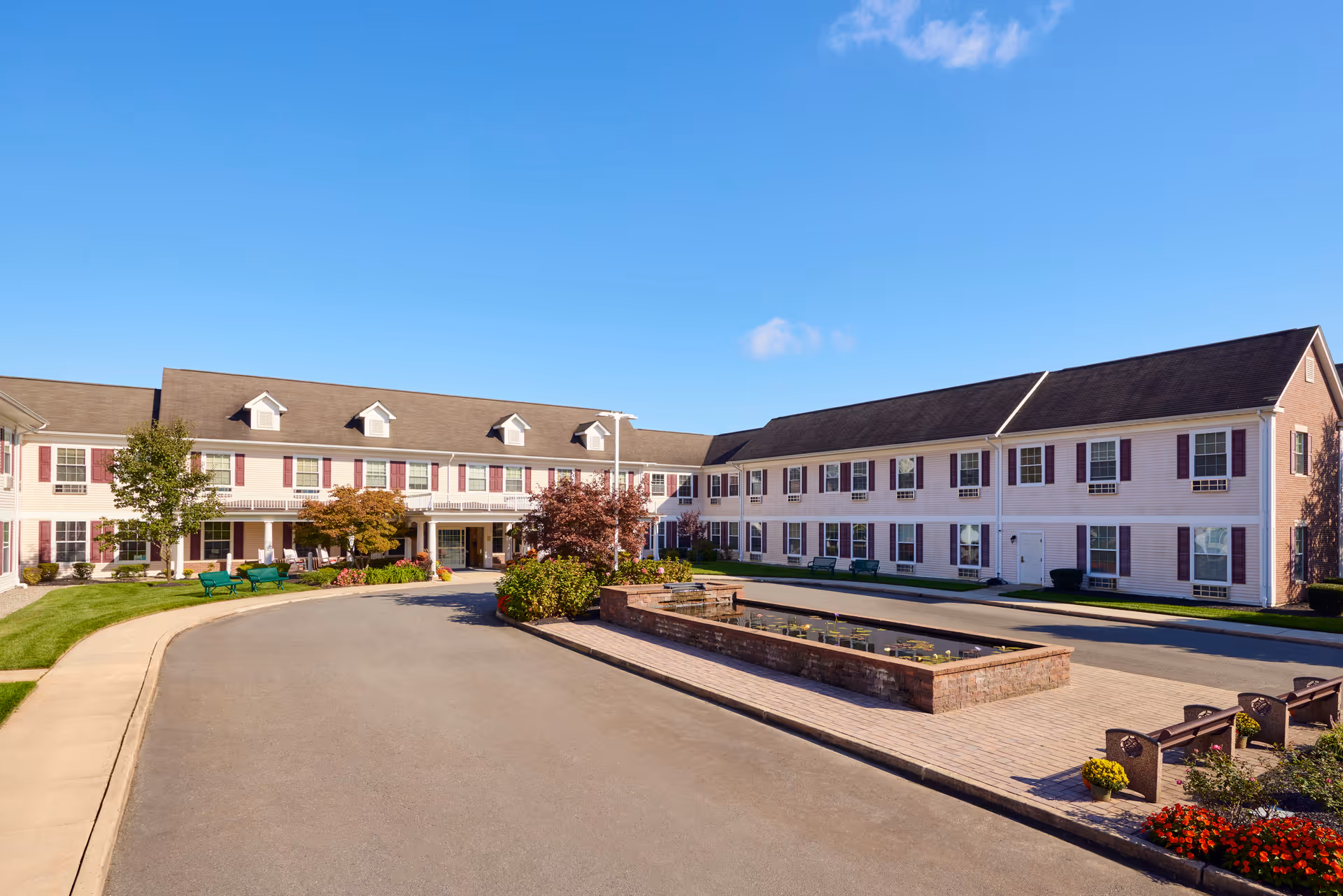 Exterior view of Brandywine Toms River by Monarch, showing a two-story building with white siding and maroon shutters under a clear blue sky. The driveway curves around a landscaped area with a rectangular water feature and benches, surrounded by green grass, trees, and flowers.