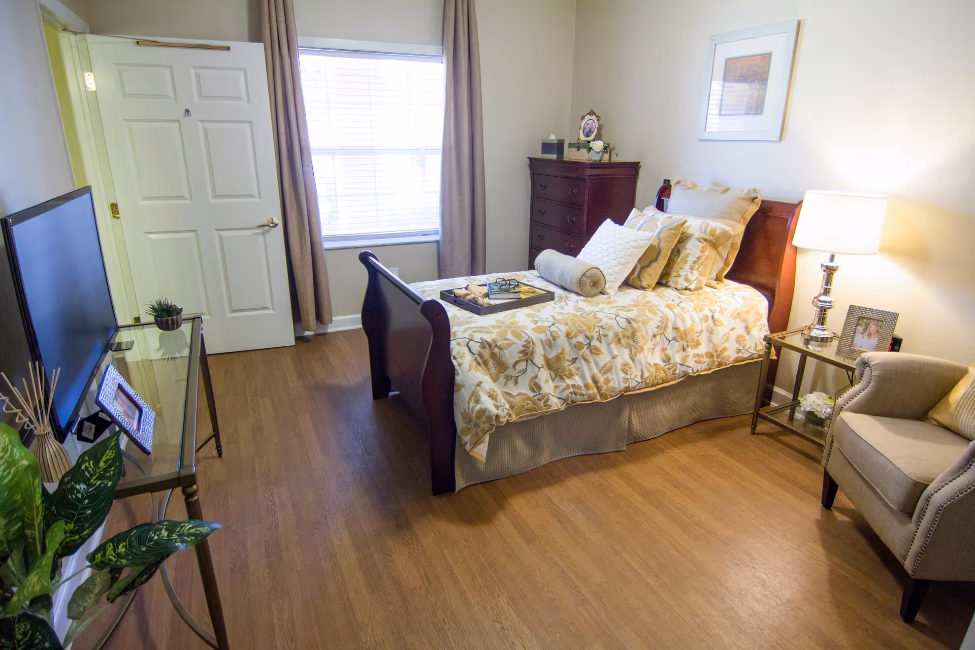 A cozy bedroom with a wooden bed featuring floral bedding and multiple pillows. There is a wooden dresser with decorative items on top, a window with beige curtains, a glass side table with a lamp and a framed photo, a beige armchair, and a flat-screen TV on a glass stand. The room has wooden flooring and light-colored walls.