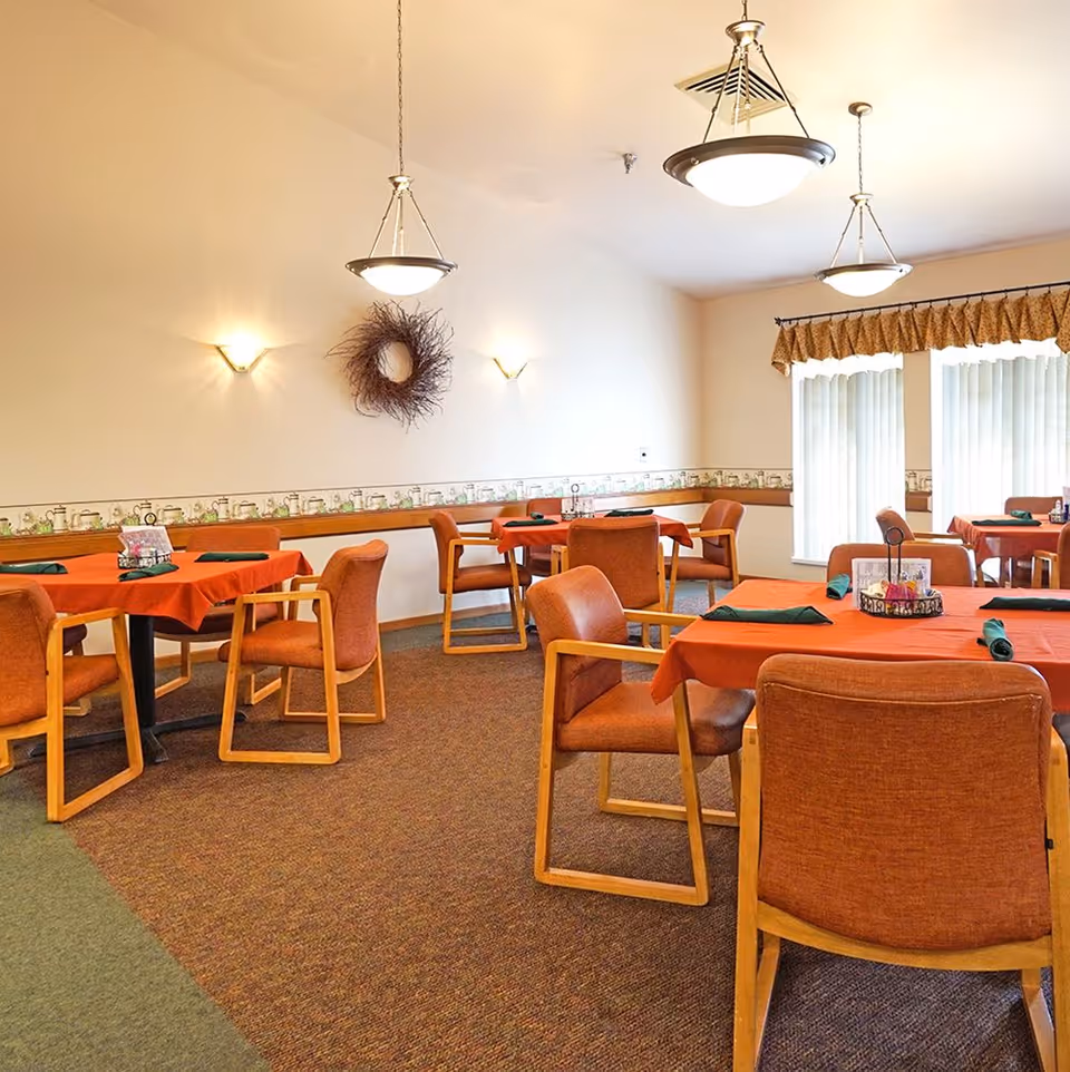 Dining room with several tables covered in red tablecloths, orange chairs, pendant lights, and large windows.