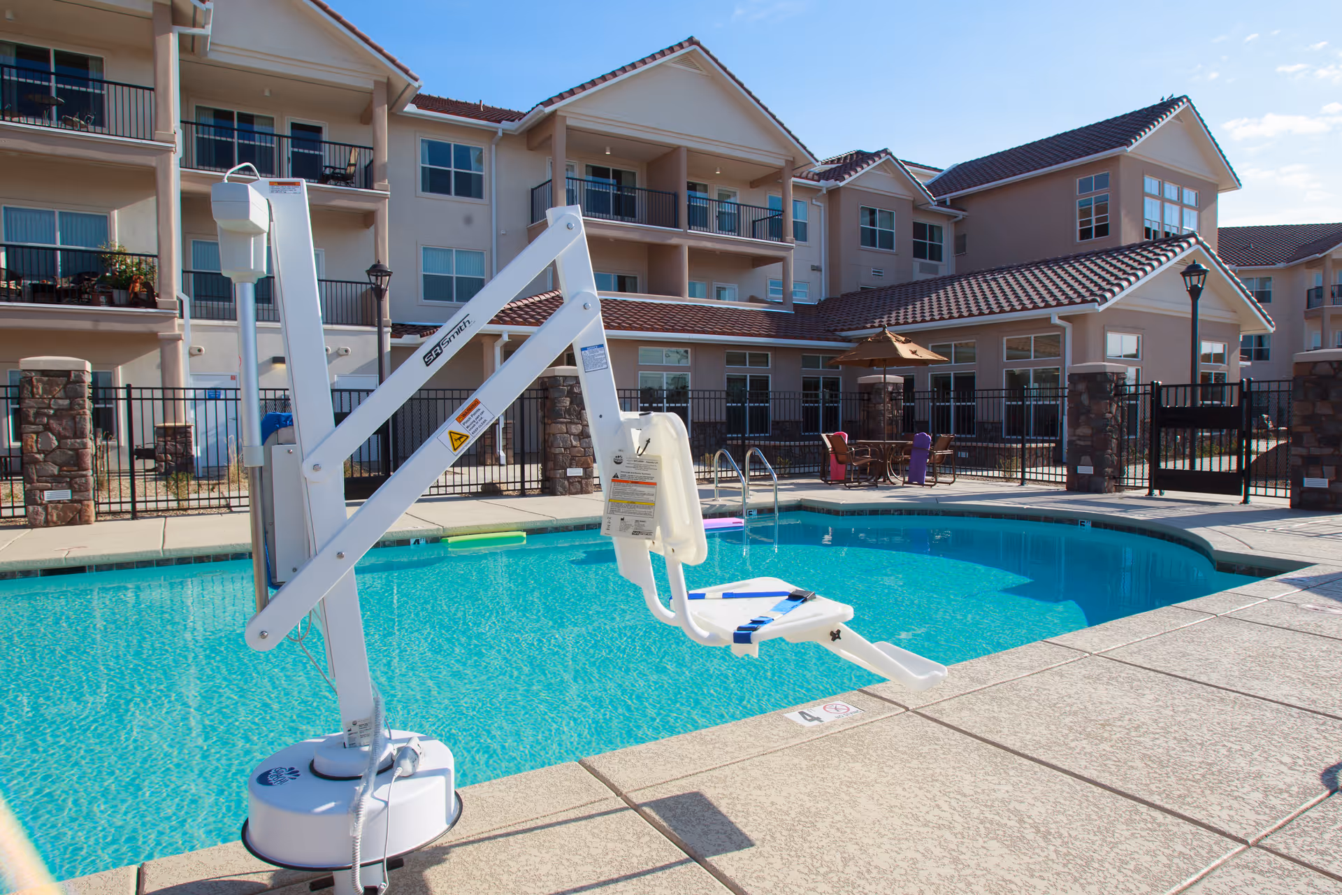 Outdoor swimming pool area at Estrella Estates Gracious Retirement Living with a pool lift for accessibility. The pool is surrounded by a concrete deck, patio furniture with umbrellas, and a multi-story residential building with balconies in the background under a clear sky.